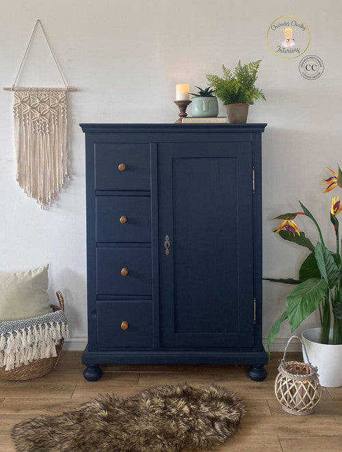 This is a tall wardrobe painted in Deep Sea Silk all-in-one mineral paint.  there are brown, round knobs to open the drawers.  On top of the wardrobe is a plant, candle stick with a white candle, a book and on top of the book is a light blue flower pot with a plant in it.  On the wall and to the left of the wardrobe is a macramé wall hanging.  Underneath that is a wicker basket with a white pillow and a blanket with tassels.  to the right of the wardrobe is a white flower pot and birds-of-paradise in it.  T