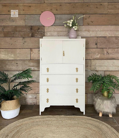 Endless Shore Silk All-In-One paint is perfect for painting kitchen cabinets, bathroom cabinets and painting furniture. White painted dressed makeover with gold handles. Staged on a wood paneled wall with a pinkish purple vase containing white flowers and a pink clock above the dresser on the wall.