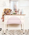 White painted dresser with white dangly drawer pulls and a natural wood top. Large half circle painted on the front drawers in light pink. Styled on a white and gray chevron rug. On top of the piece there is a large wire basket with a yellow and white striped blanket hanging out, a book with a flower bouquet placed on top, and three colorful wine bottles. The piece is in front of a white wood paneled wall with a picture of a horse head on in the middle above the dresser.