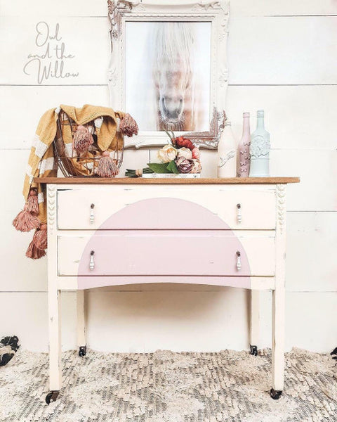 White painted dresser with white dangly drawer pulls and a natural wood top. Large half circle painted on the front drawers in light pink. Styled on a white and gray chevron rug. On top of the piece there is a large wire basket with a yellow and white striped blanket hanging out, a book with a flower bouquet placed on top, and three colorful wine bottles. The piece is in front of a white wood paneled wall with a picture of a horse head on in the middle above the dresser.