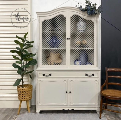 Endless Shore Silk All-In-One paint is perfect for painting kitchen cabinets, bathroom cabinets and painting furniture. White painted armoire with black handles. Styled next to a woven planter containing faux greenery. Wooden chair placed on the right of the armoire.
