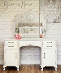 Endless Shore Silk All-In-One paint is perfect for painting kitchen cabinets, bathroom cabinets and painting furniture. White painted vanity with a large mirror and metal handles. Pink roses placed on top of the vanity. Staged on a wood floor in front of a white brick wall and a picture of a white chandelier hung on the wall.