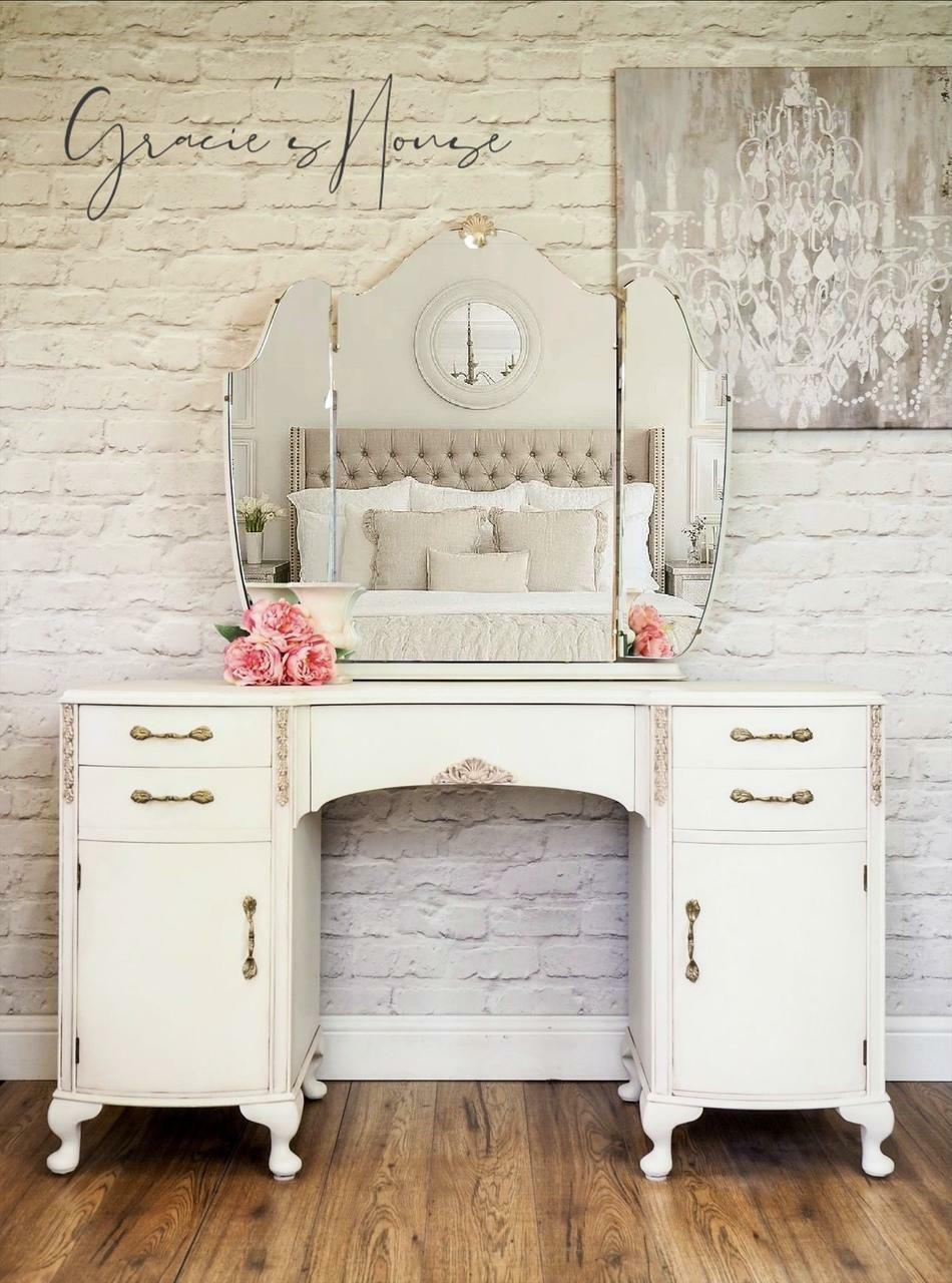 Endless Shore Silk All-In-One paint is perfect for painting kitchen cabinets, bathroom cabinets and painting furniture. White painted vanity with a large mirror and metal handles. Pink roses placed on top of the vanity. Staged on a wood floor in front of a white brick wall and a picture of a white chandelier hung on the wall.