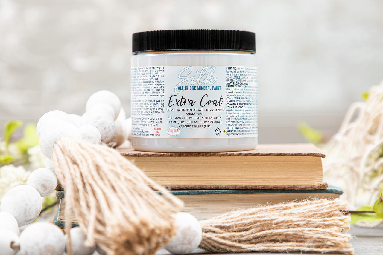 Silk All-In-One Mineral Paint Extra Coat. 16 oz. jar of Extra Coat placed on top of 2 books with wooden beads with yarn on the ends placed next to the jar and books.