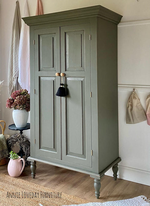Hampton Olive Silk All-In-One paint is perfect for painting kitchen cabinets, bathroom cabinets and painting furniture.
A wardrobe painted in Hampton Olive Silk All-In-One paint, staged with flower vases and blankets hanging on the wall besides the piece.