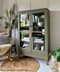 Hampton Olive Silk All-In-One paint is perfect for painting kitchen cabinets, bathroom cabinets and painting furniture.
Glass display case painted in Hampton Olive Silk All-In-One paint staged with boho decor around the piece like a brown woven rug, potted houseplants, and a chair with pillows and a fur blanket.