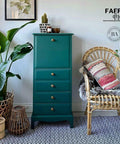 Midnight Green Silk All-In-One paint is perfect for painting kitchen cabinets, bathroom cabinets and painting furniture. Dark green Silk all In one painted tall dresser. Staged with some wicker potted plants a nd a boho style chair with pillows and blankets next to it. On top has a potted cactus, a wood bowl and candle.