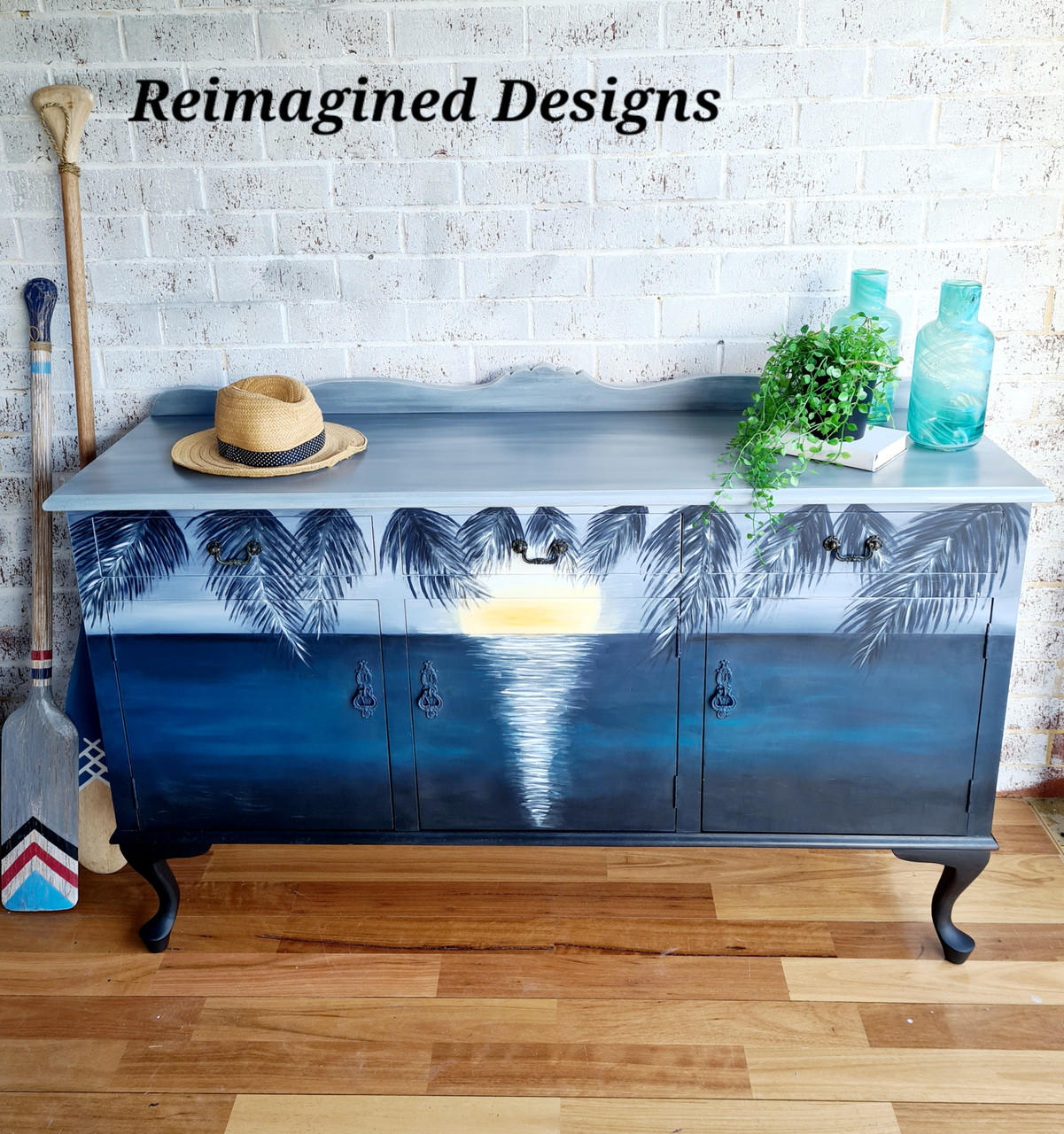 Nautical Silk All-In-One paint is perfect for painting kitchen cabinets, bathroom cabinets and painting furniture. Long hutch with a painted sunset and ocean painted with different shades of blue chalk paint. Staged with glass vases, a plant and a garden hat.
