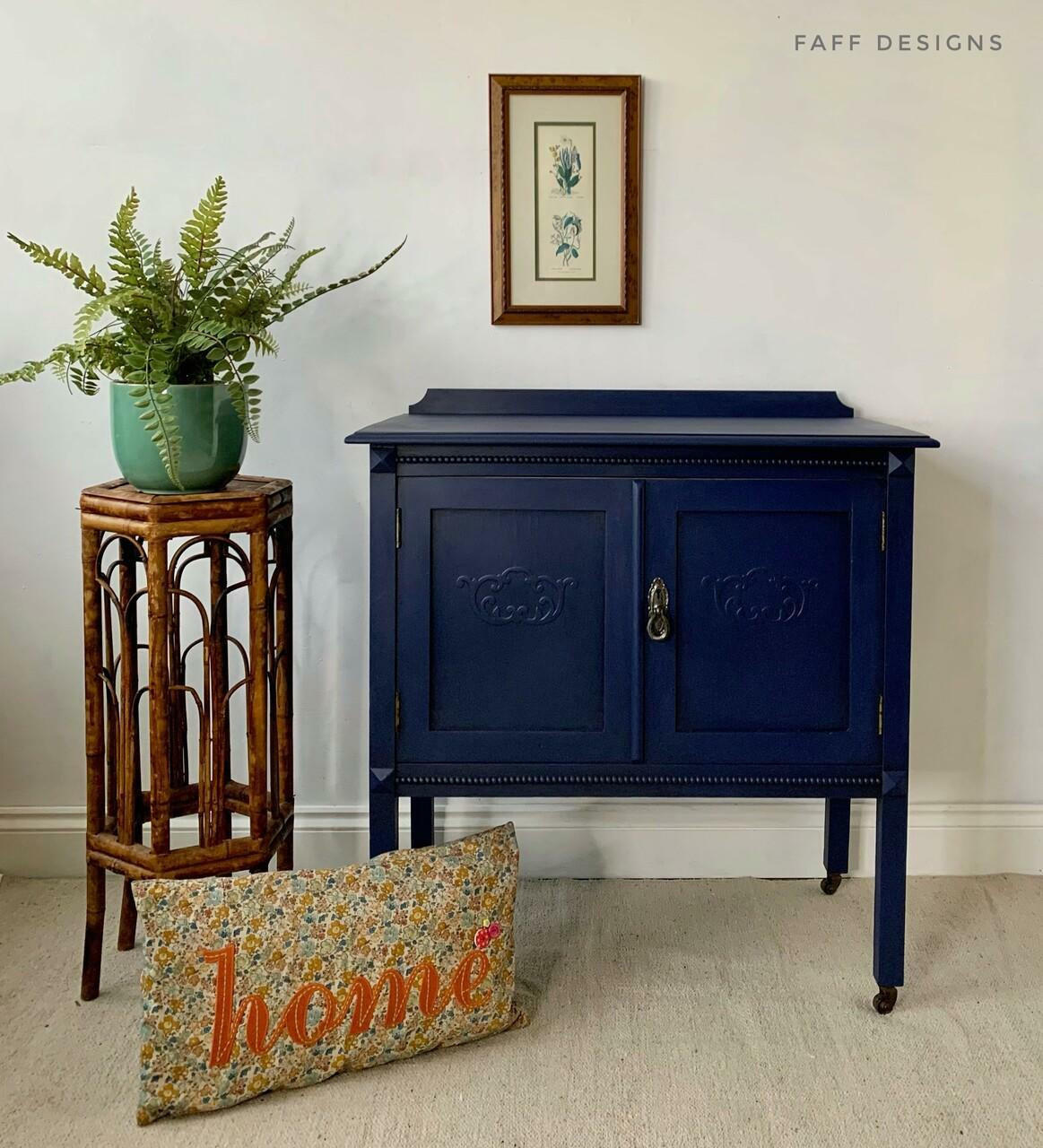 Nautical Silk All-In-One paint is perfect for painting kitchen cabinets, bathroom cabinets and painting furniture. Dark blue chalk painted cabinet with gold hardware. Staged next to a potted plant with a pillow.