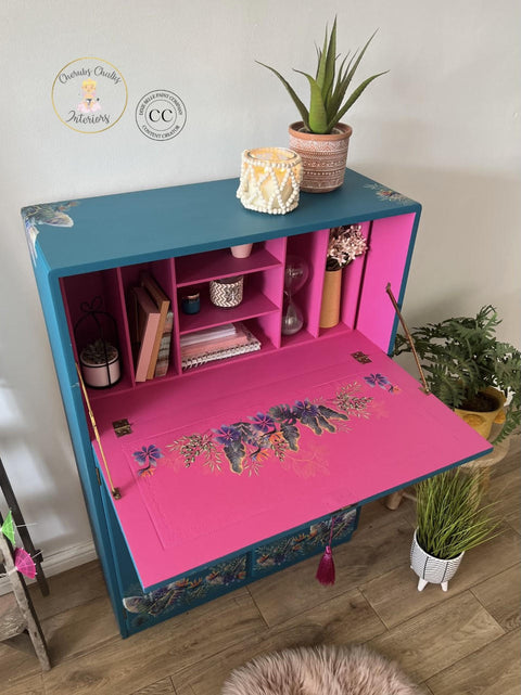 Oasis Silk All-In-One paint is perfect for painting kitchen cabinets, bathroom cabinets and painting furniture. Blue chalk painted secretary desk with pink chalk painted inside. Tropical leaves rub on transfer on the front and inside the desk. Staged with books, and candles and a plant.