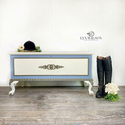 Quiet Cove Silk All-In-One paint is perfect for painting kitchen cabinets, bathroom cabinets and painting furniture! A chest that has a white top and white front but the trim is this medium-gray color and with gray around the bottom trim. This chest has a wooden applique on the front painted in gray and is staged against a white wall with a pair of boots to the right, and a cap and book on the top of the chest.