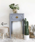 Silk All-In-One Mineral Paint Quiet Cove Silk Paint is perfect for painting kitchen cabinets, bathroom cabinets and painting furniture! A medium-gray colored nightstand with one drawer, golden hardware, and long legs. This nightstand is staged with a cactus to the right, wooden basket with a blanket, a short stool to the left and a potted plant on top.