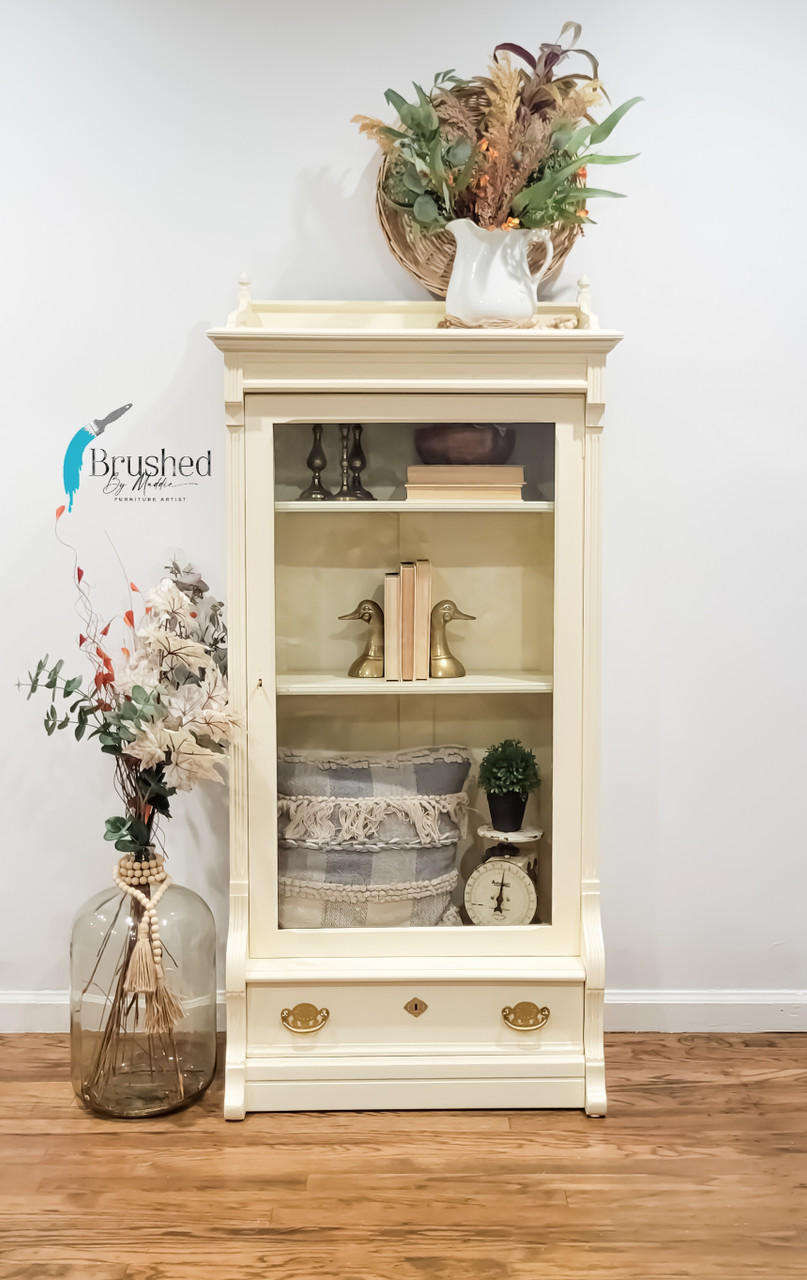 Sun Kissed Silk All-In-One paint is perfect for painting kitchen cabinets, bathroom cabinets and painting furniture.
Small display cabinet painted in Sun Kissed Silk All-In-One paint with one drawer at the bottom with gold hardware. Staged with niknaks, books and baskets inside the cabinet, besides piece is a large glass jug with flowers and branches.