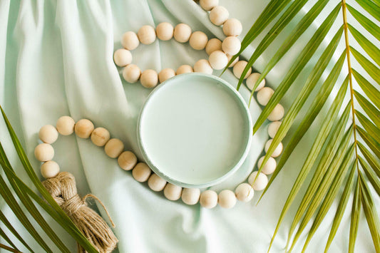 Tide Pool Silk All-In-One paint is perfect for painting kitchen cabinets, bathroom cabinets and painting furniture.  An open jar of Tide Pool Silk All-in-One paint on a blue-green colored piece of cloth, with ivory beads and palm leaves around.