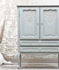 Wharf Silk All-In-One paint that is perfect for painting kitchen cabinets, bathroom cabinets and painting furniture.  A gray chalk painted armoire sits on a light colored wooden floor, in front of a brown and white wall.  The front of the armoire has light colored embellishments in the center of each door.  o the left of the armoire is a white vase with brown foliage.