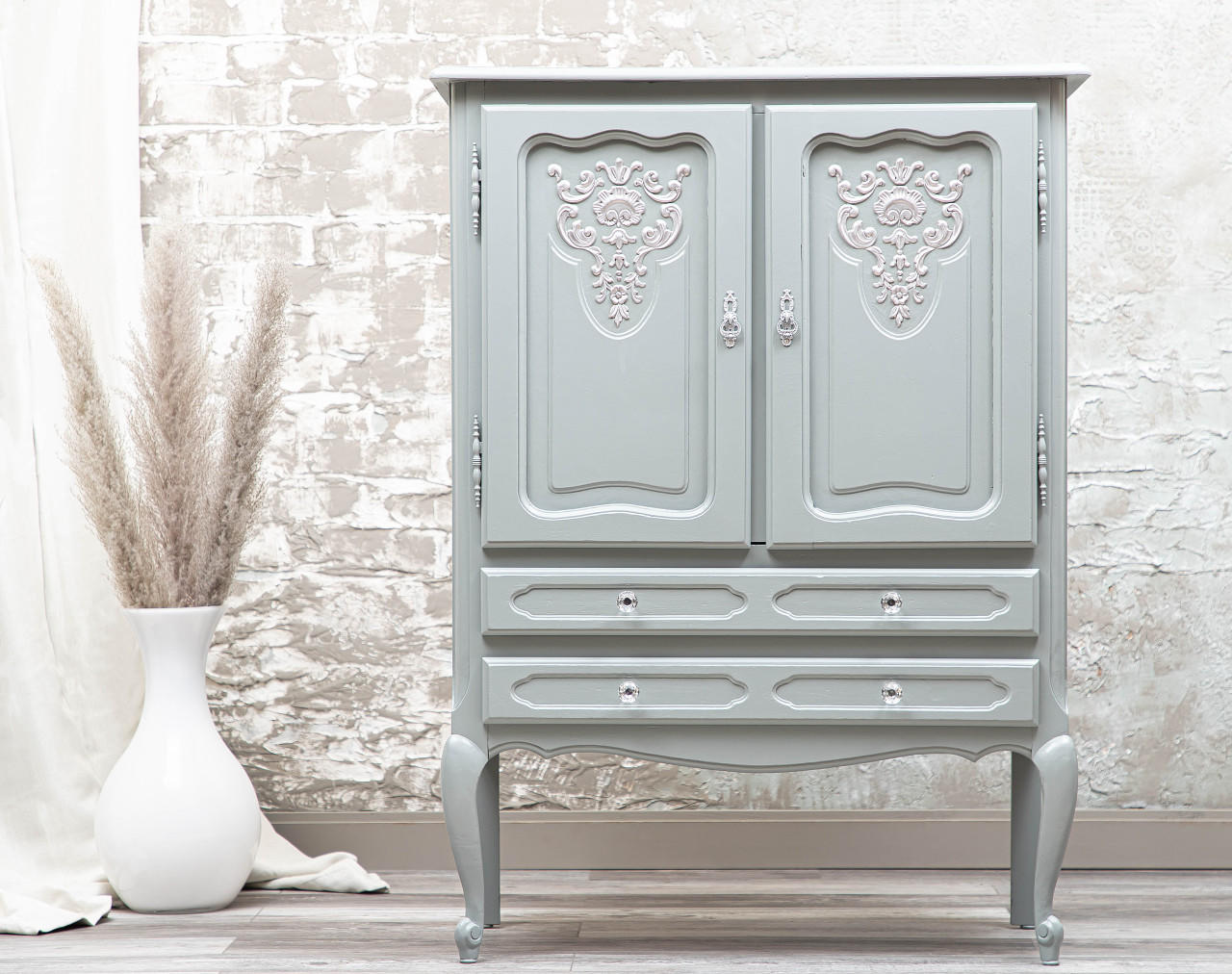 Wharf Silk All-In-One paint that is perfect for painting kitchen cabinets, bathroom cabinets and painting furniture.  A gray chalk painted armoire sits on a light colored wooden floor, in front of a brown and white wall.  The front of the armoire has light colored embellishments in the center of each door.  o the left of the armoire is a white vase with brown foliage.