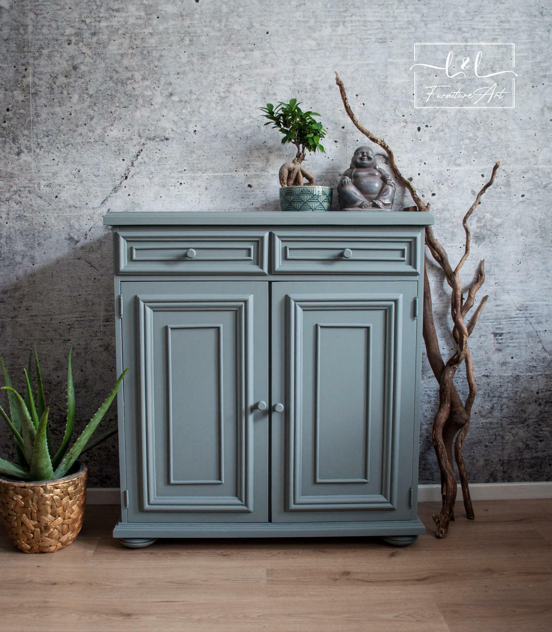 Wharf Silk All-In-One paint that is perfect for painting kitchen cabinets, bathroom cabinets and painting furniture.  A gray chalk painted cabinet sitting on a light brown wooden floor, in front of a gray wall.  A green aloe plant sits in a gold pot on the floor to the left of the cabinet and brown wooden sticks stand up against the wall to the right of the cabinet.  A buddha statue and a small green plant in a blue pot sit on top of the cabinet.