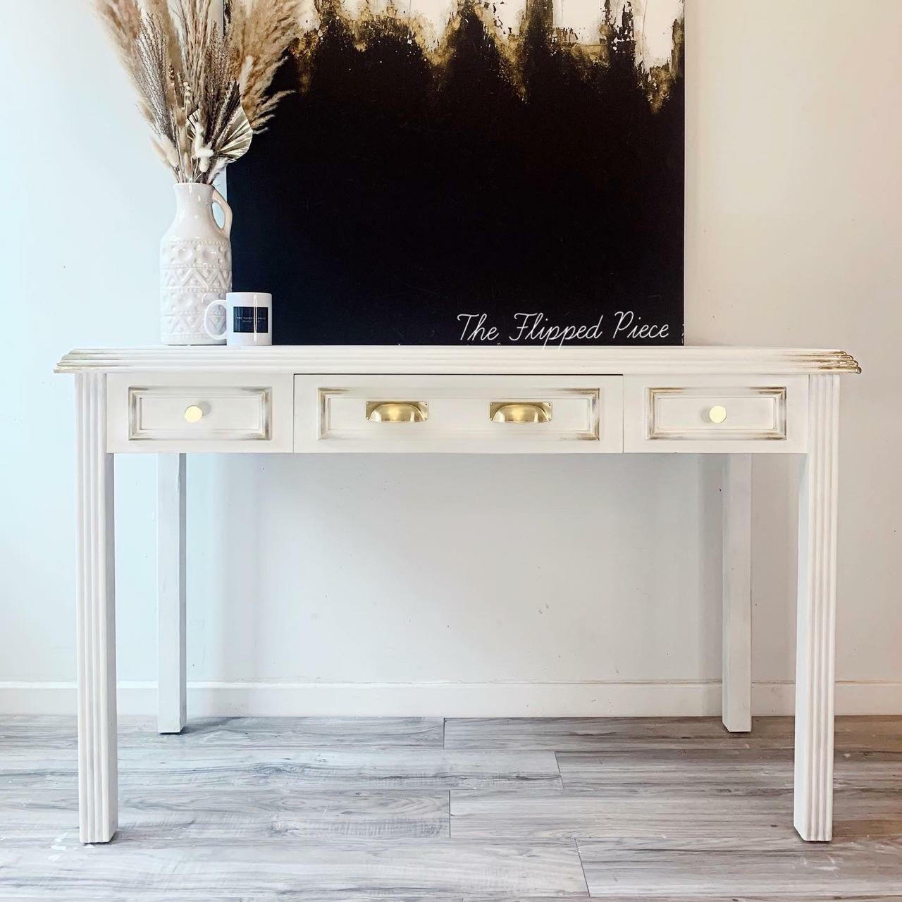 Whitecap Silk All-In-One paint is perfect for painting kitchen cabinets, bathroom cabinets and painting furniture.  A white chalk painted hall table with gold distressed corners and gold handles on the drawers sits on a light brown wooden floor, in front of a white wall.  A black, white and gold painting sits on top of the desk, along with a white and black coffee cup and a white vase holding wheat.