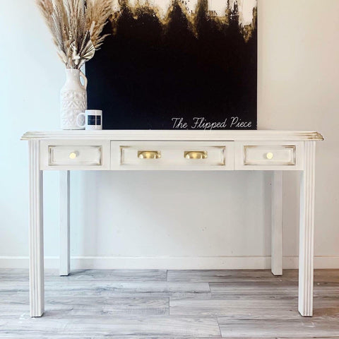 Whitecap Silk All-In-One paint is perfect for painting kitchen cabinets, bathroom cabinets and painting furniture.  A white chalk painted hall table with gold distressed corners and gold handles on the drawers sits on a light brown wooden floor, in front of a white wall.  A black, white and gold painting sits on top of the desk, along with a white and black coffee cup and a white vase holding wheat.