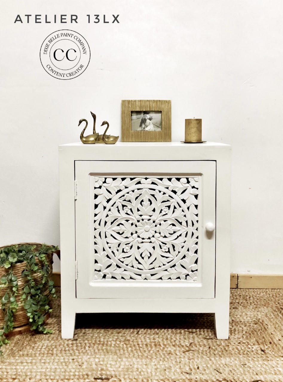 Whitecap Silk All-In-One paint is perfect for painting kitchen cabinets, bathroom cabinets and painting furniture.  A white chalk painted corner table with a door sits on a brown woven rug, in front of a white wall.  On the floor to the left of the night stand is a green plant inside a woven basket.  Gold figurines, a gold picture frame, and a gold candle sit on top of the night stand.