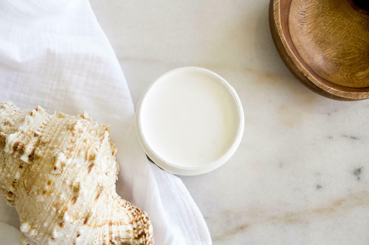 Whitecap Silk All-In-One paint is perfect for painting kitchen cabinets, bathroom cabinets and painting furniture.  A top view of an open jar of Whitecap Silk All-In-One paint sitting on a white marble table.  A white linen cloth sits to the left of the jar with a white and brown conch shell sitting on top of it.  And a circular piece of brown wood sits to the right of the jar of paint.