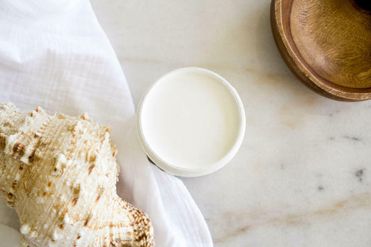Whitecap Silk All-In-One paint is perfect for painting kitchen cabinets, bathroom cabinets and painting furniture.  A top view of an open jar of Whitecap Silk All-In-One paint sitting on a white marble table.  A white linen cloth sits to the left of the jar with a white and brown conch shell sitting on top of it.  And a circular piece of brown wood sits to the right of the jar of paint.