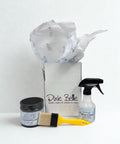 A Dixie Belle gift box with tissue paper, a jar of silk mineral paint, a yellow paintbrush, and a spray bottle of Pristine Clean displayed in front on a white background.