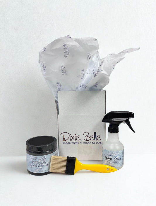 A Dixie Belle gift box with tissue paper, a jar of silk mineral paint, a yellow paintbrush, and a spray bottle of Pristine Clean displayed in front on a white background.