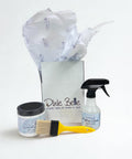 A Dixie Belle gift box with tissue paper, a jar of silk all-in-one mineral paint, a bottle of surface cleaner, and a yellow paintbrush displayed in front on a white background.