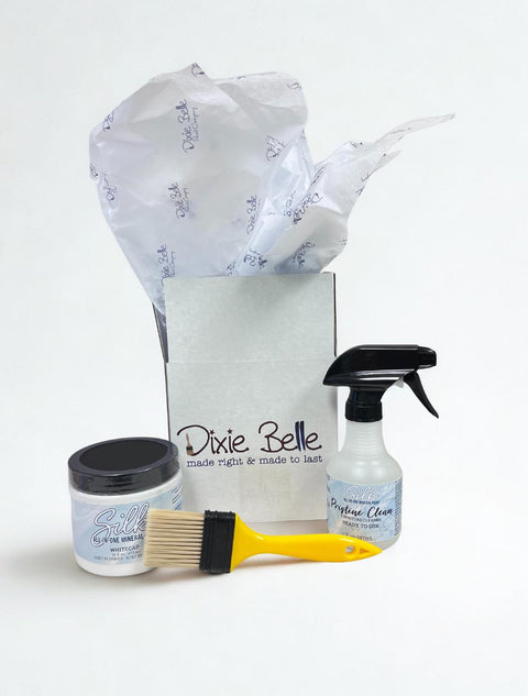 A Dixie Belle gift box with tissue paper, a jar of silk all-in-one mineral paint, a bottle of surface cleaner, and a yellow paintbrush displayed in front on a white background.