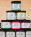 A stack of Silk all-in-one mineral paint jars in various colors, including green, pink, yellow, and brown. Each jar has a label detailing the paint's color, name, and brand information. The background is a soft, neutral tone.