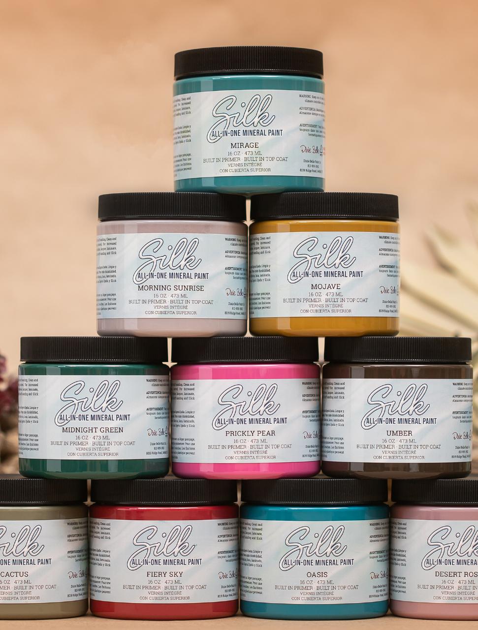 A stack of Silk all-in-one mineral paint jars in various colors, including green, pink, yellow, and brown. Each jar has a label detailing the paint's color, name, and brand information. The background is a soft, neutral tone.