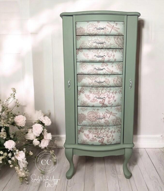 A vintage-style cabinet painted in green with six drawers. Each drawer features a floral and dragonfly pattern design in subtle colors. Two silver handles are on the side. The background includes white flowers on the left and a white wall.