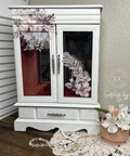 A white jewelry armoire decorated with floral designs stands on a wooden surface. It features a drawer at the bottom. A lace doily, a glass bead necklace, and a vase with white flowers sit in front of the armoire.