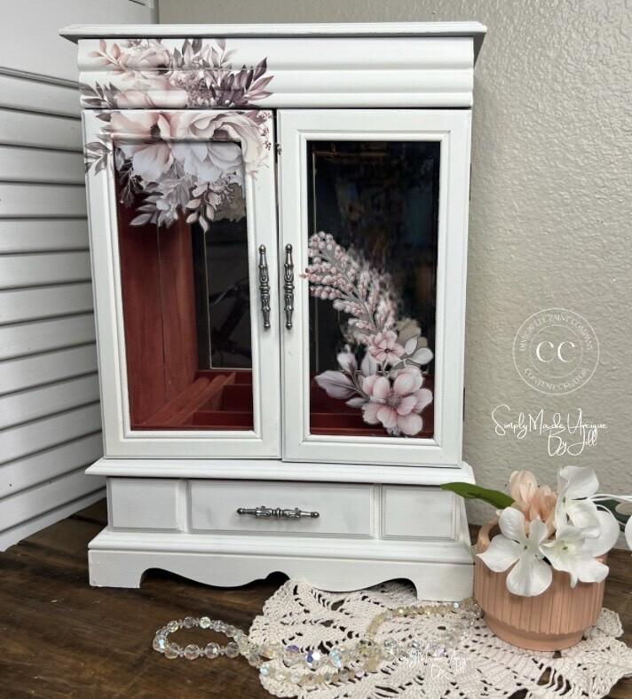A white jewelry armoire decorated with floral designs stands on a wooden surface. It features a drawer at the bottom. A lace doily, a glass bead necklace, and a vase with white flowers sit in front of the armoire.