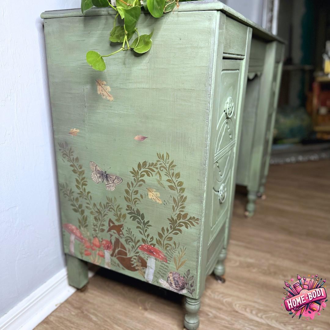 A vintage green desk adorned with intricate leaf, butterfly, and mushroom designs on its side. A vibrant green plant rests atop the desk. A "HOME·BODI" logo is placed on the lower right corner, and the desk stands on a wooden floor.