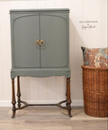 Bluish gray painted armoire makeover with gold circular pulls and wooden legs. Staged next to a large wicker basket with a multicolor pillow inside.