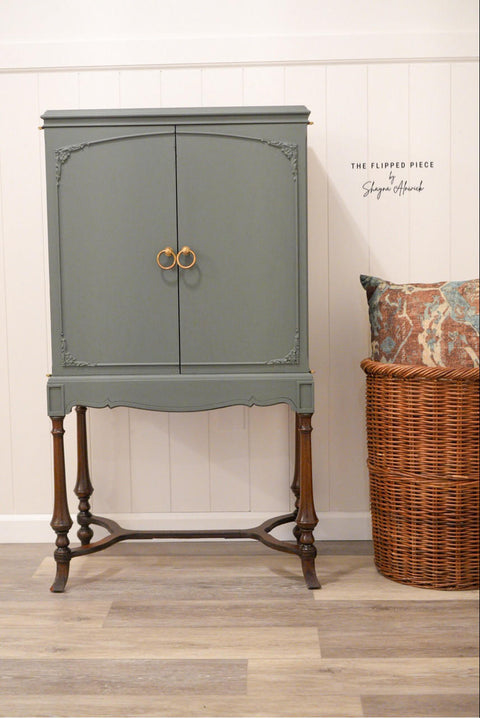 Bluish gray painted armoire makeover with gold circular pulls and wooden legs. Staged next to a large wicker basket with a multicolor pillow inside.