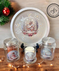 A festive display showing a round tray with a Christmas scene of a snowy house. Three glass jars, adorned with winter images of a snowman, pine tree, and reindeer, are in front. A candle burns inside one jar. A small red ornament and greenery are in the background.