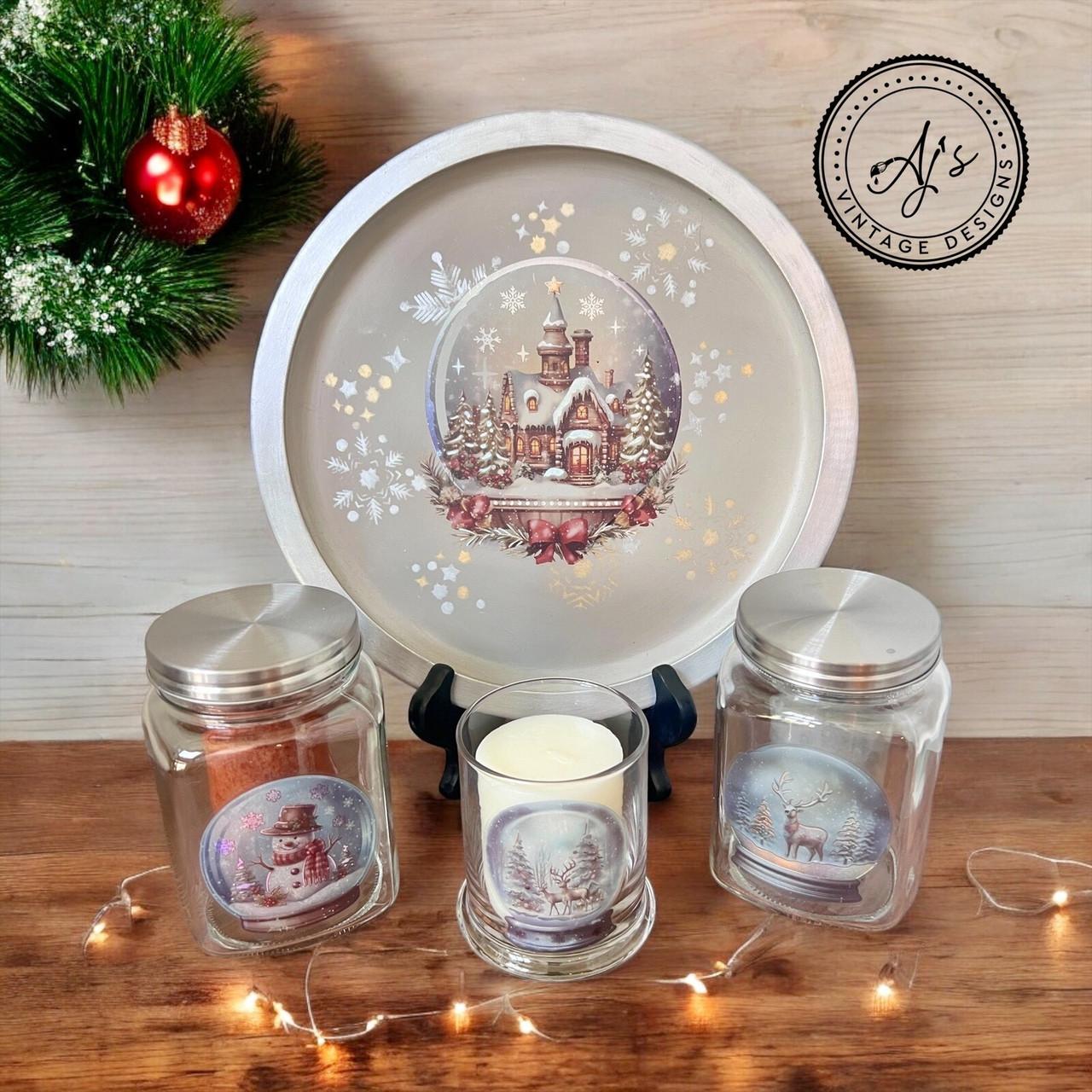A festive display showing a round tray with a Christmas scene of a snowy house. Three glass jars, adorned with winter images of a snowman, pine tree, and reindeer, are in front. A candle burns inside one jar. A small red ornament and greenery are in the background.