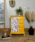 A bright yellow dresser with four drawers sits against a gray shiplap wall. The dresser's front has a partial floral motif featuring pastel flowers. Various decor items, including plants, a woven basket lantern, and a black vase with pampas grass, surround it.