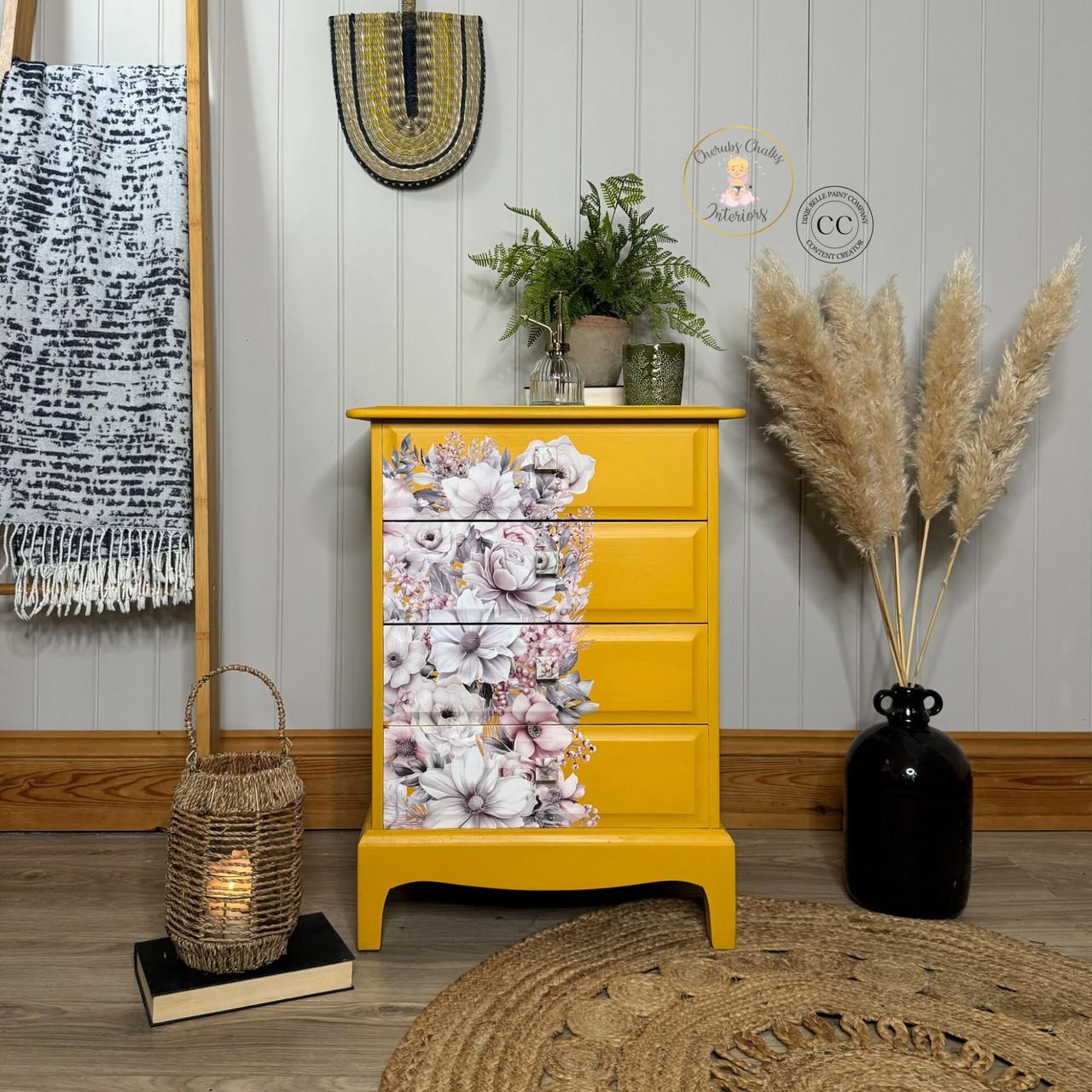 A bright yellow dresser with four drawers sits against a gray shiplap wall. The dresser's front has a partial floral motif featuring pastel flowers. Various decor items, including plants, a woven basket lantern, and a black vase with pampas grass, surround it.