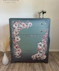 A repurposed dresser painted in gradient teal with floral designs in pink and white on the front. The dresser has three drawers with silver handles and a decorative central panel. It is placed against a wall, with a vase of dried grass and white flowers on top.