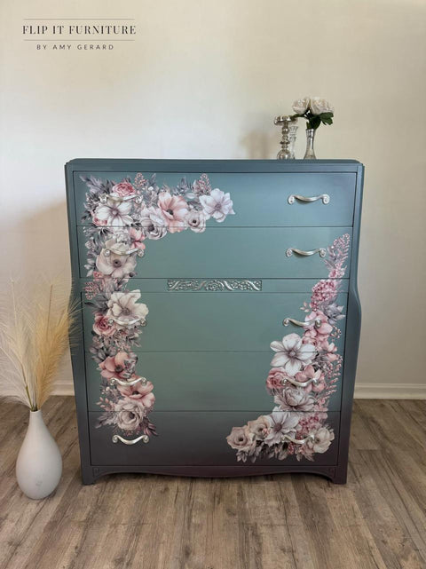 A repurposed dresser painted in gradient teal with floral designs in pink and white on the front. The dresser has three drawers with silver handles and a decorative central panel. It is placed against a wall, with a vase of dried grass and white flowers on top.