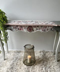 A decorative table with floral details and scalloped edges stands on a plush, grey rug. A leafy plant drapes over its left side. Below, a large glass candle holder with a lit candle sits on the floor, adding a warm, cozy ambiance to the scene.