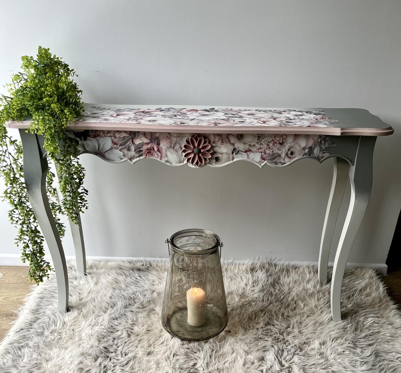 A decorative table with floral details and scalloped edges stands on a plush, grey rug. A leafy plant drapes over its left side. Below, a large glass candle holder with a lit candle sits on the floor, adding a warm, cozy ambiance to the scene.