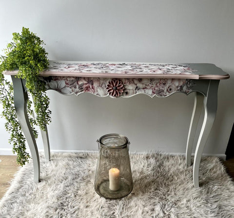 A decorative table with floral details and scalloped edges stands on a plush, grey rug. A leafy plant drapes over its left side. Below, a large glass candle holder with a lit candle sits on the floor, adding a warm, cozy ambiance to the scene.