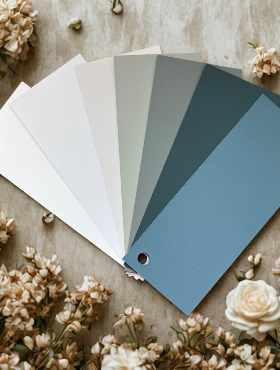 A fan of six paint color swatches ranging from off-white to blue-green is displayed on a beige surface, surrounded by small white flowers.