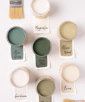 An arrangement of seven open paint cans, each labeled with a name and a paintbrush nearby. Paint colors include white, green, beige, and olive shades, with labels: Magnolia, Blue Grass, Brian Patch, Olive Wood, Gardenia, Thicket, and Ecru.