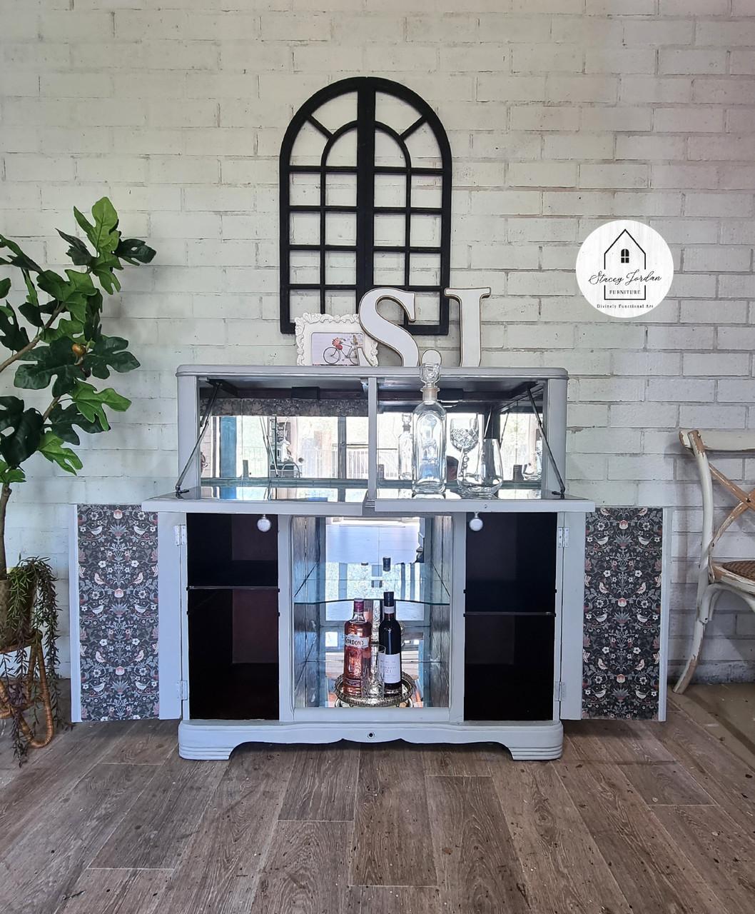 A gray cabinet with bottles inside is against a white brick wall. It has a decorative mirror on top, flanked by a potted plant. A semicircular window decor and a round sign are above. Flooring is wood-patterned.